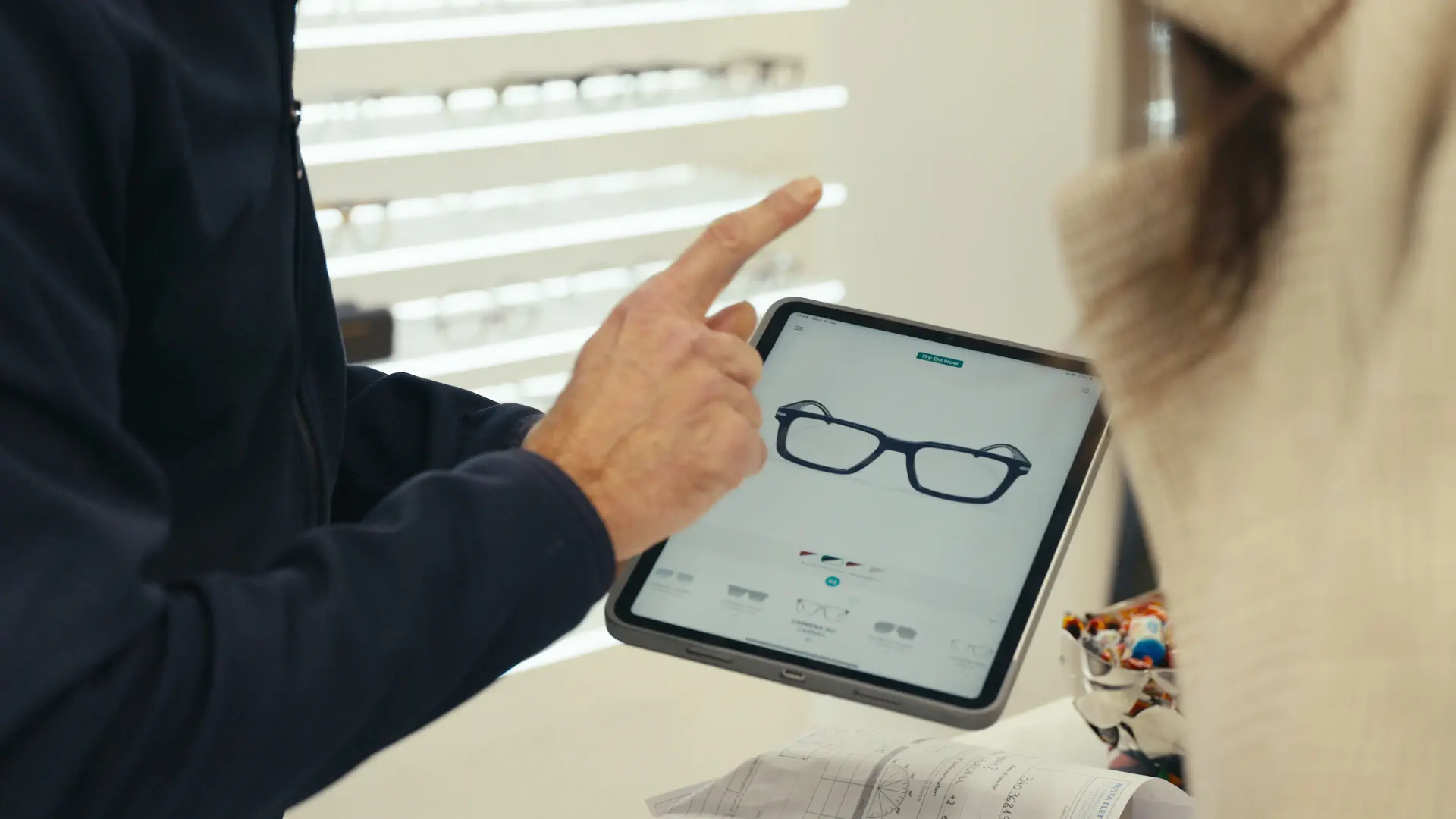 In einem Geschäft zeigt ein Optiker auf ein Tablet: Der 3D Viewer stellt eine blaue Fassung mit ihren Farbvarianten dar.