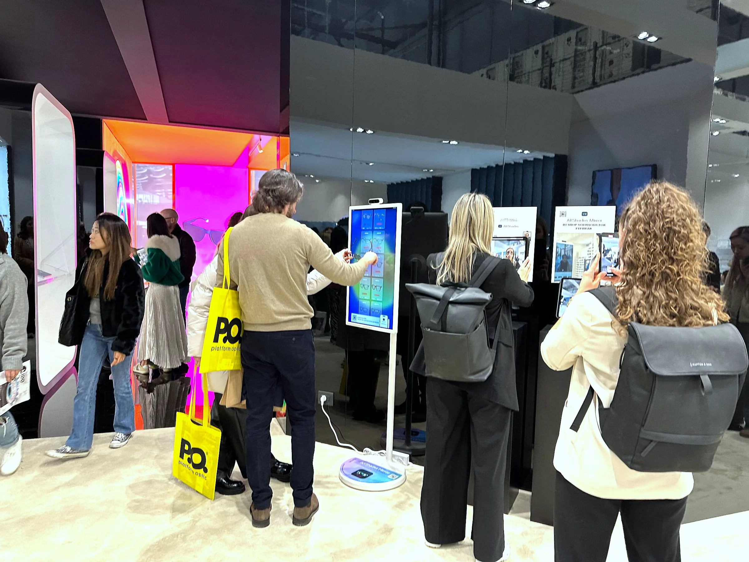 An einem Messestand blättert ein Besucher auf einem digitalen Spiegel im Hochformat durch die Kollektion, um ihn herum stehen weitere Personen.