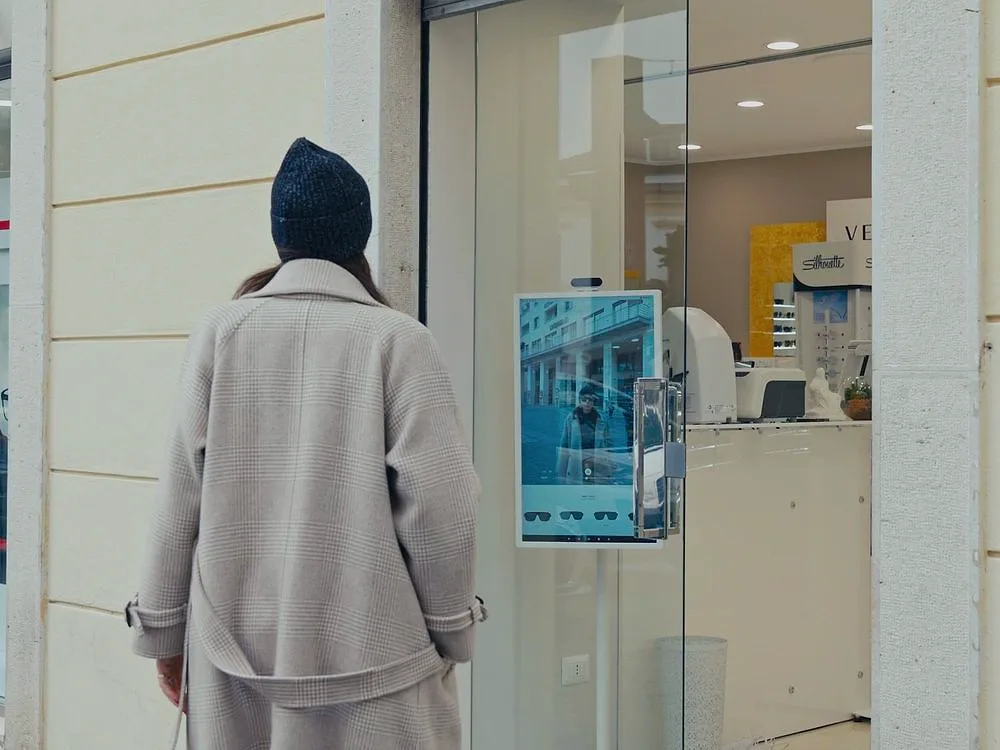 Eine Passantin bleibt vor dem Schaufenster eines Optikers stehen, wo ein digitaler Spiegel sie von der Straße aus ins Bild nimmt.