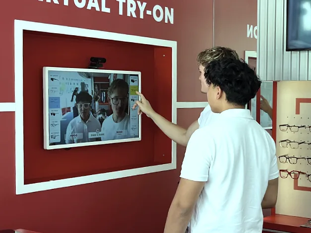 Zwei junge Leute vor einem Bildschirm, der in eine rote Wand mit dem Schriftzug Virtual Try-On eingelassen ist. Sie sehen sich darin gemeinsam, jeder mit seiner eigenen Brille.