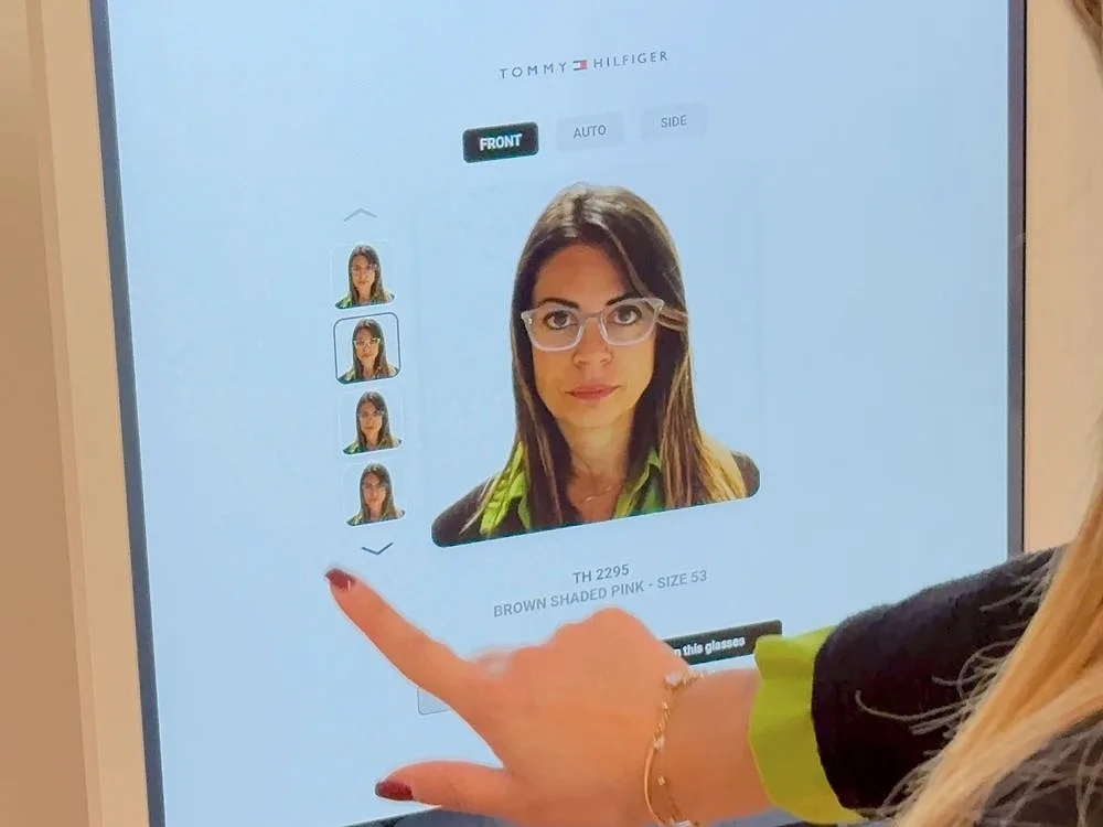 Eine Kundin berührt den digitalen Spiegel. Auf dem Bildschirm ist ihr Gesicht mit Brille zu sehen, daneben dasselbe Gesicht mit anderen Fassungen.