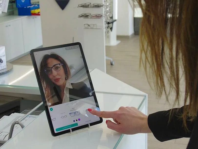 Auf der Theke eines Optikers steht ein Tablet. Eine Kundin sieht sich mit der Brille im Gesicht und wählt auf dem Bildschirm die Fassung aus.