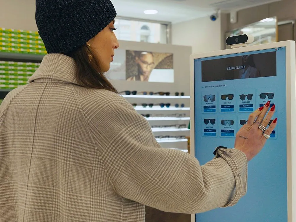 In einem Optikgeschäft wählt eine Kundin Sonnenbrillen aus, indem sie auf dem Bildschirm des digitalen Spiegels die Fassungen antippt.