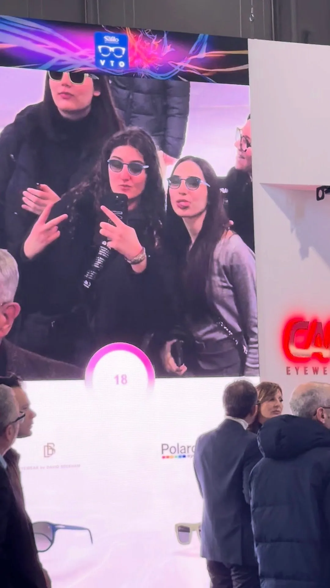 Auf einer Messe sehen sich drei junge Frauen auf einem riesigen Bildschirm mit virtuellen Sonnenbrillen und posieren für das Foto.