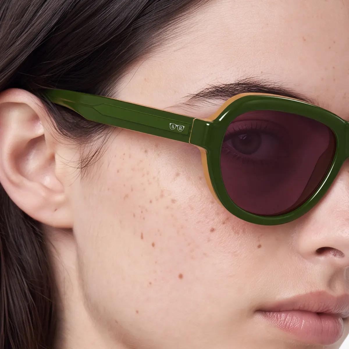Close-up of a dark-haired woman wearing Rover sunglasses: olive-green acetate with visible reflections, brown lenses and a small metal logo on the temple.